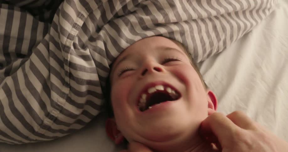 A child lies in bed laughing uncontrollably while being tickled by an adults hands