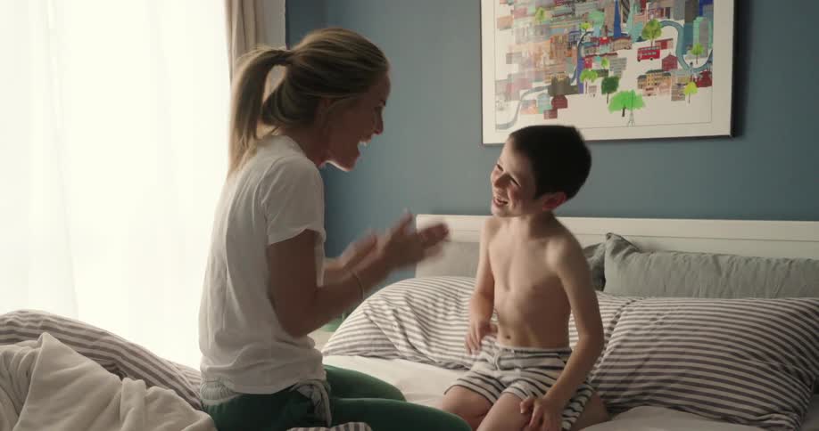 A woman and a young boy are sitting on a bed playing a handclapping game in a cozy sunlit bedroom