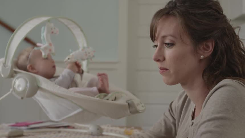 A mom sits while her baby plays in a chair behind her