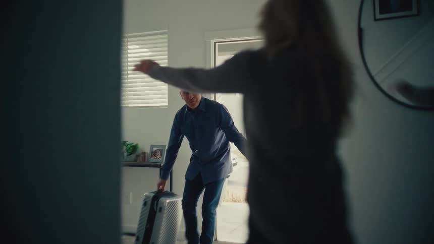 Wife happily running into husbands arms as we walks in the door carrying luggage
