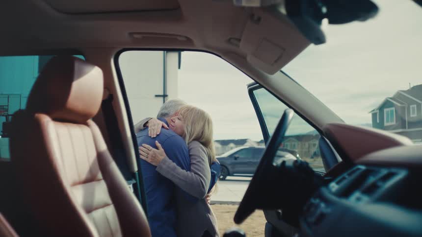 From inside a car with an open door a woman embraces a man in a heartfelt hug both mature adults sharing a sentimental moment in a neighborhood during the day