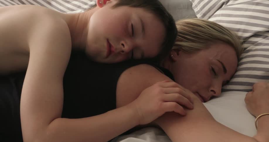 A son lies on his mothers back resting for a nap on the bed while gently caressing her shoulder in a moment of relaxation