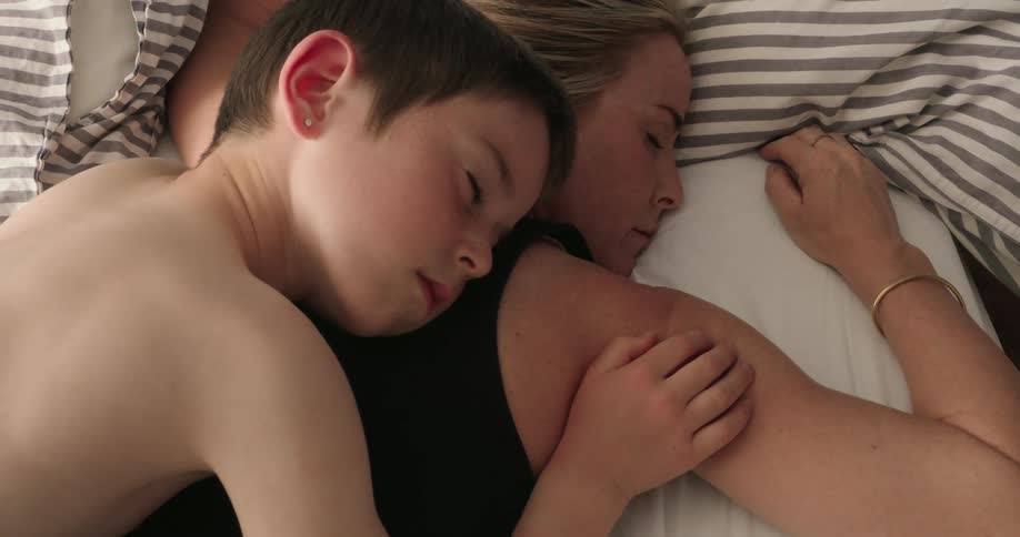 A young boy peacefully sleeps nestled against a woman in a quiet intimate moment on a bed