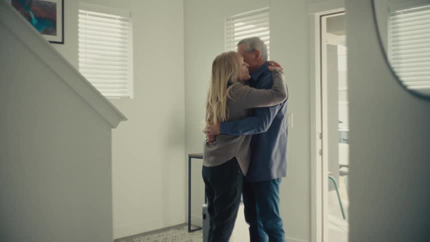 An adult couple shares a warm hug and smile during an emotional sendoff inside a bright modern home