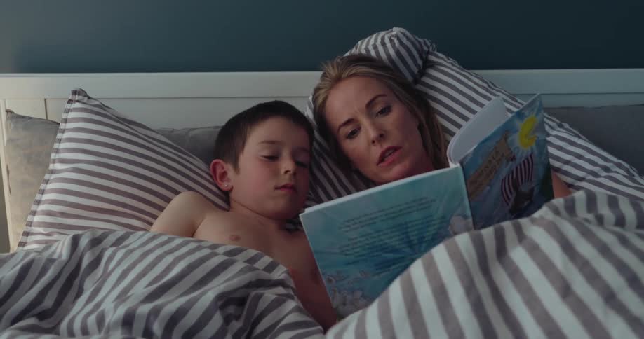 A woman and young boy read a childrens book together in bed sharing a quiet intimate moment