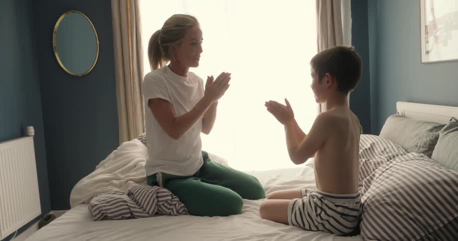 A mother and her son sit on the bed playing by clapping high fives as she smiles and teaches him at midday
