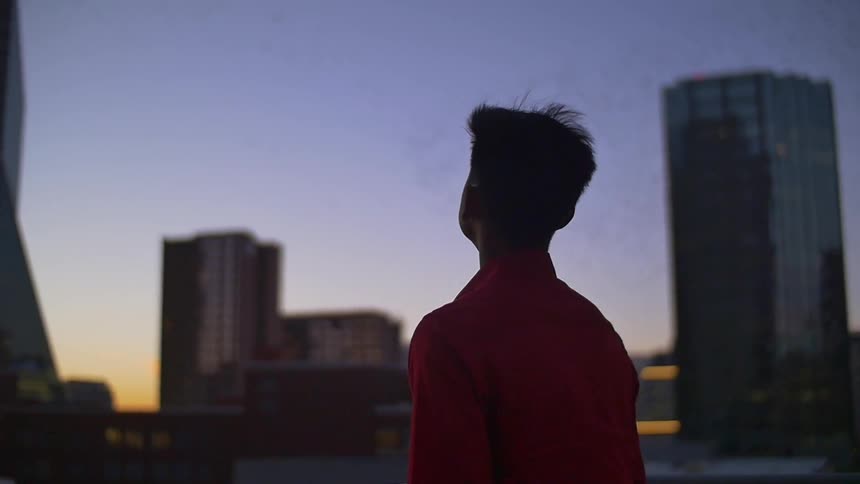 A young asian male stands looking up at a tall building and the city at dusk