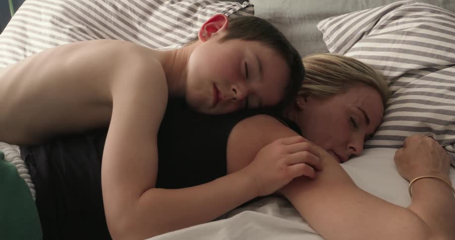 A son lies on his mothers back resting for a nap on the bed while gently caressing her shoulder in a moment of relaxation