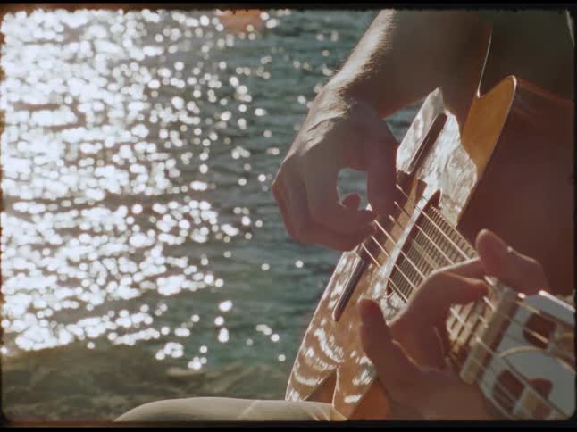 Person playing guitar by the sea with bright reflections on the water in the background
