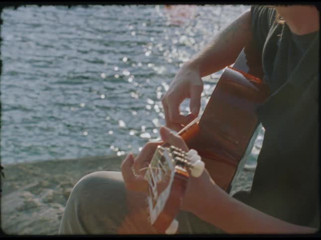 A man plays an acoustic guitar by the shimmering water captured in a peaceful sunlit moment