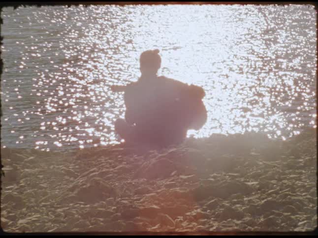 An adult is sitting on a river bank and playing the guitar at sunset