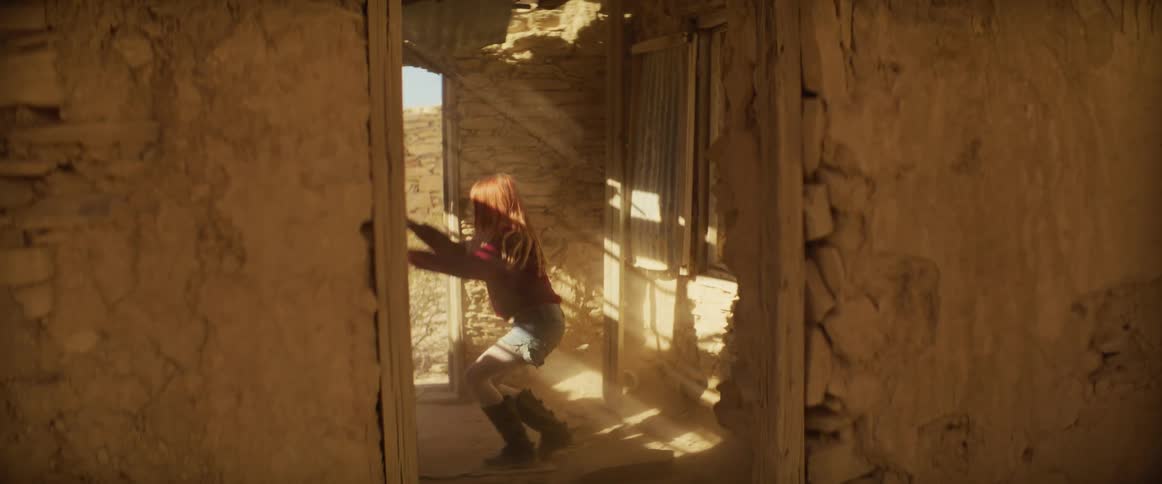 A woman jumps out of a crumbling adobe building into the sunlight and stands facing the open desert