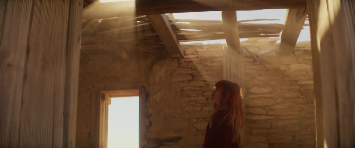 A woman walks through sunlit ruins of a stone building light streaming through the broken roof