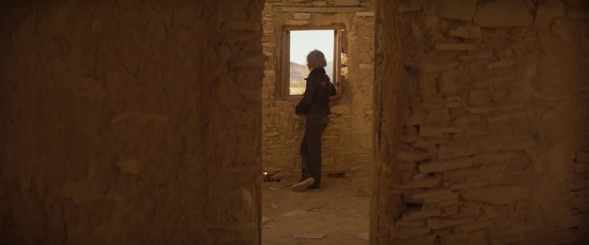 A young boy stands in a dusty crumbling stone room glancing back at the camera while facing an open window