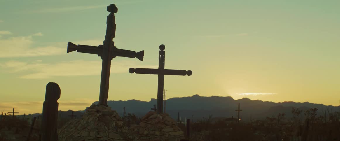 Two wooden crosses stand atop stonecovered graves in a desert cemetery at sunset silhouetted by distant mountains