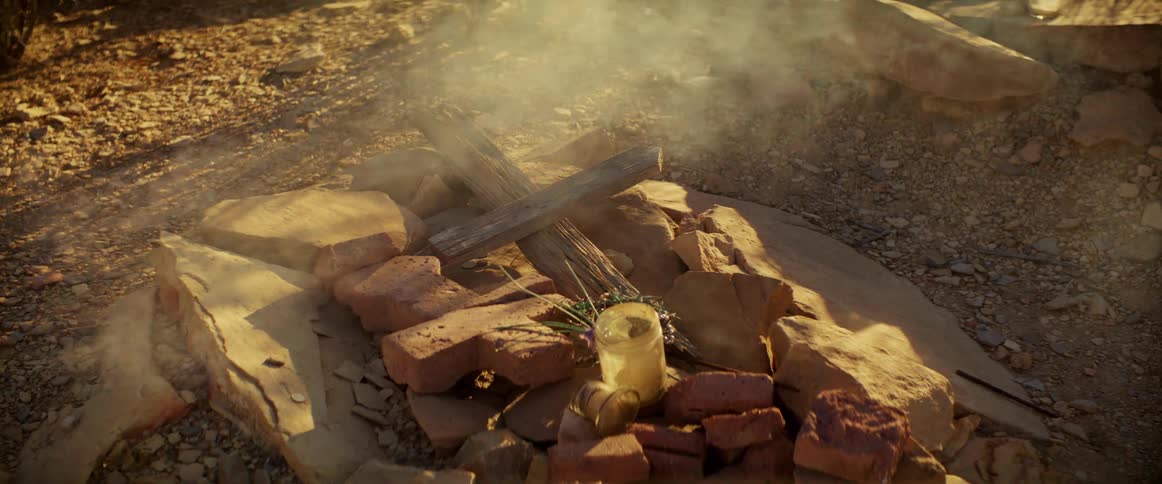 A makeshift grave marked by a wooden cross and surrounded by stones in a desert landscape with a candle and flowers placed at the base