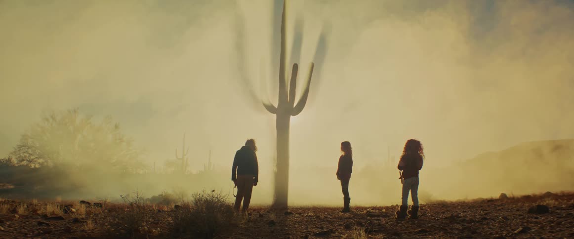 Three kids stand in a dusty desert at dusk facing a towering cactus silhouetted in the haze