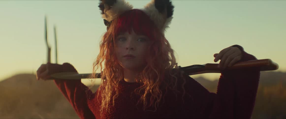 A girl with red hair and animal ears stands in desert at dusk holding shovel across shoulders