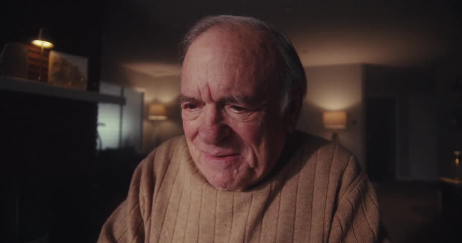 A closeup of an elderly mans face captures his expression of happy surprise as he thinks in front of a screen in a softly lit room