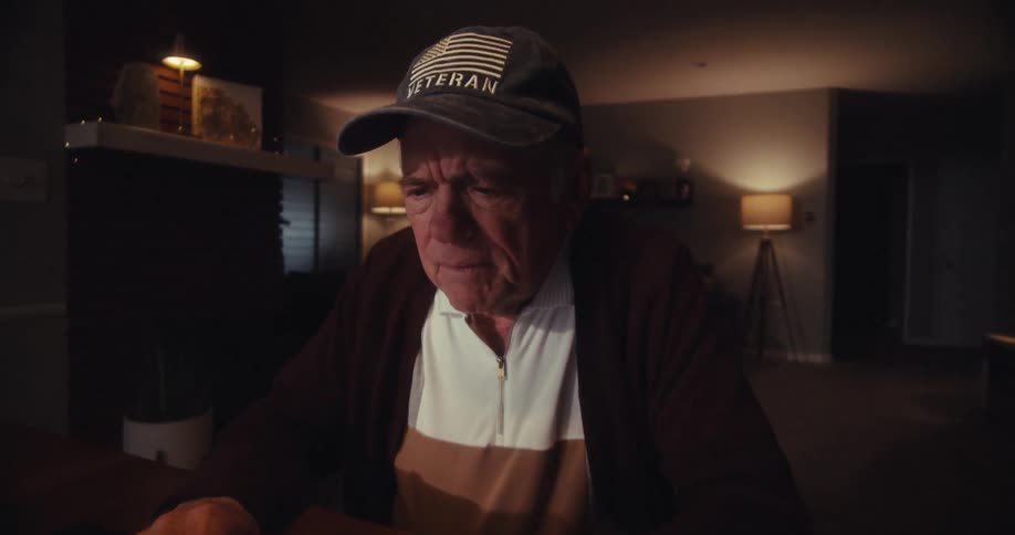 A closeup of an elderly man wearing a veteran baseball hat sitting in a dimly lit living room focused intently on the laptop he is working on