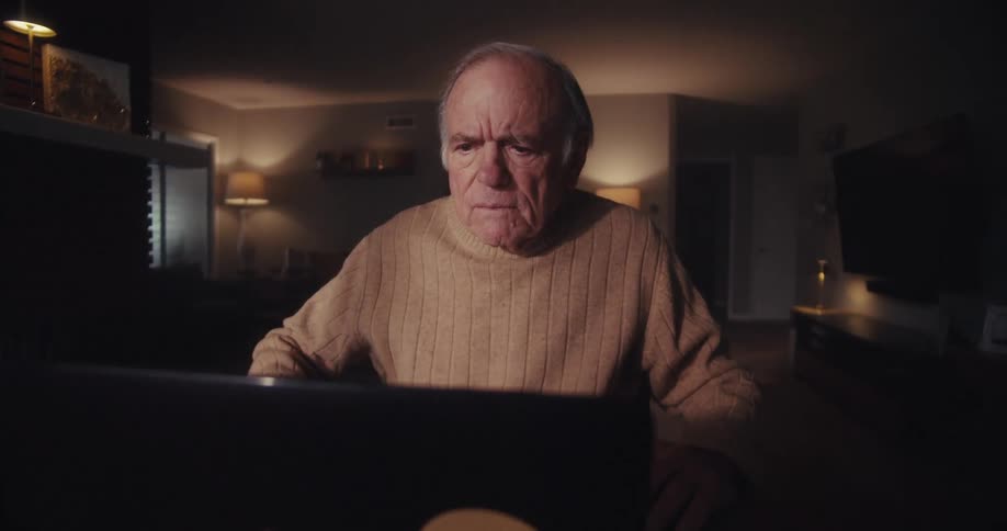 Elderly man focused on his laptop while sitting at home at night reacts with surprise and pleasure