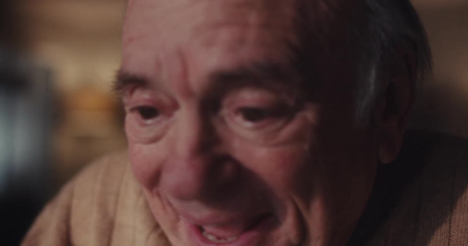 A closeup of an elderly mans face captures his expression of happy surprise as he looks at a screen in a softly lit room