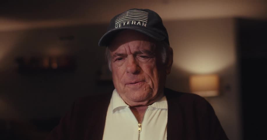 An older man wearing a veteran baseball cap smiles and looks down at a laptop in a dimly lit room