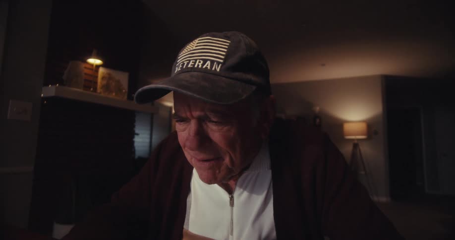 An elderly man wearing a dark cap with an american flag and the word veteran smiles and laughs while watching a screen in his home