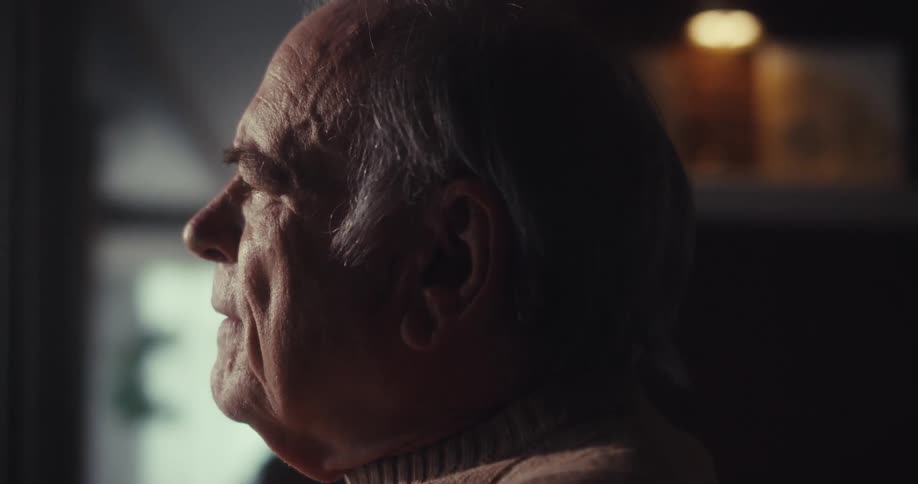 Elderly man stares into space before focusing downward while sitting indoors