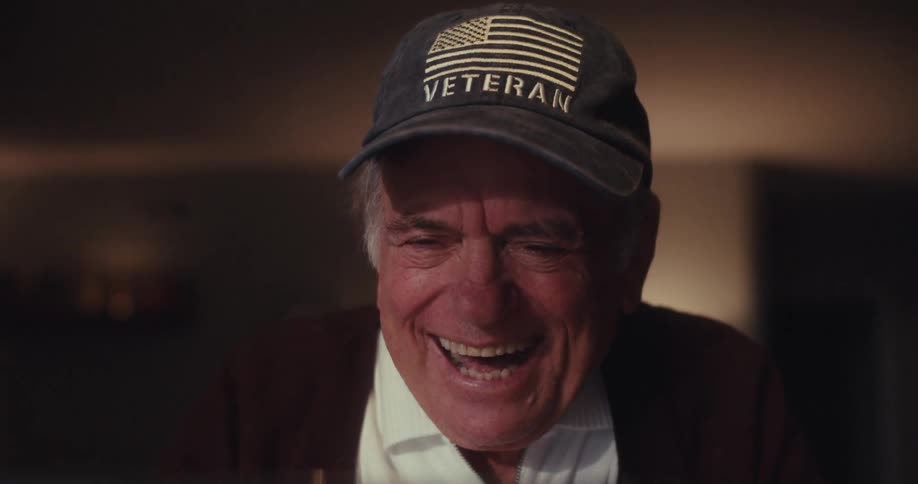 An elderly man wearing a dark cap with an american flag and the word veteran smiles and laughs while watching a screen in his home