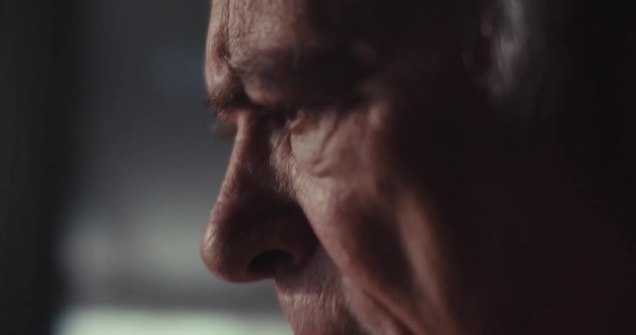 A closeup of an elderly man sitting in a dimly lit living room deep in thought and fully focused on his task