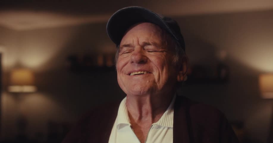 An elderly man relaxes at home in a dimly lit living room contentedly watching a screen and stretching