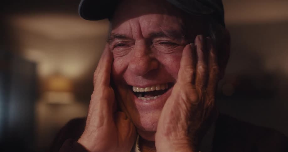 An elderly man wearing a dark cap with an american flag and the word veteran smiles and laughs while watching a screen in his home