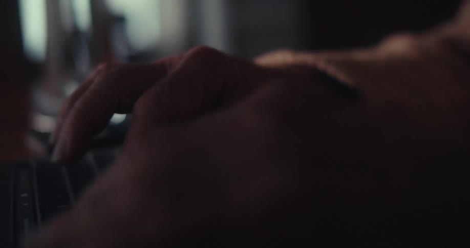An elderly man sits at a table in a dimly lit living room focused as he works on his laptop