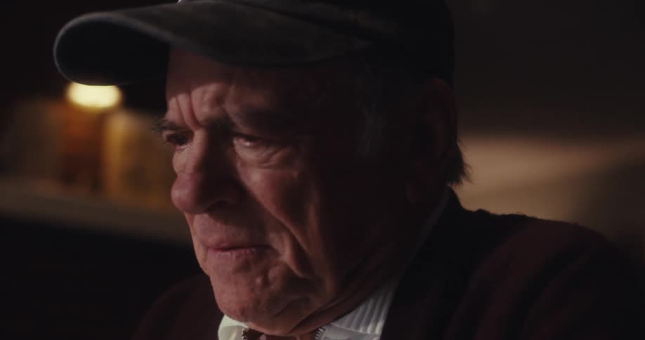 An elderly man wearing a dark cap with an american flag and the word veteran embroidered on it sits in a dimly lit room his expression heavy with disappointment and sadness
