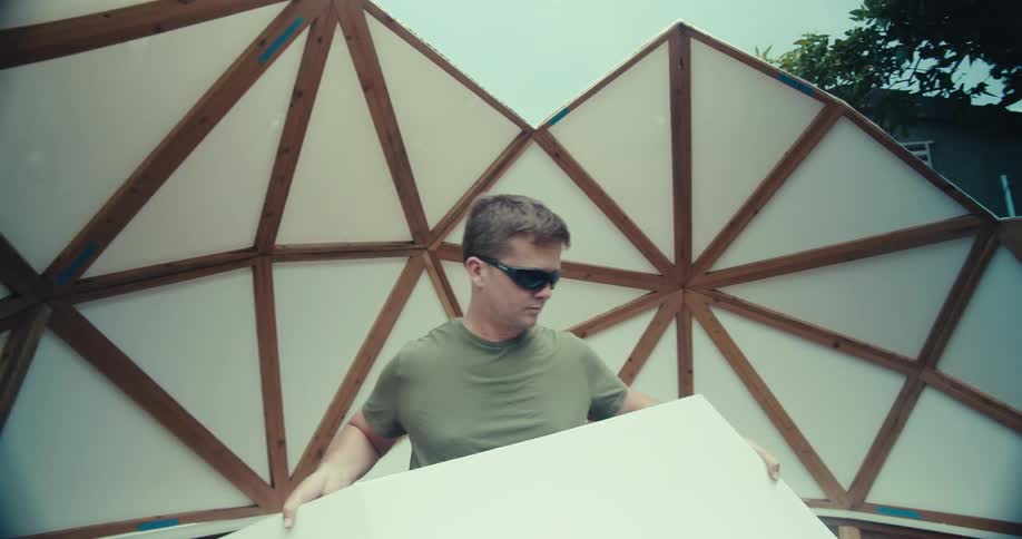 A person wearing sunglasses is working on the construction of a geodesic dome structure adjusting plastic coverings