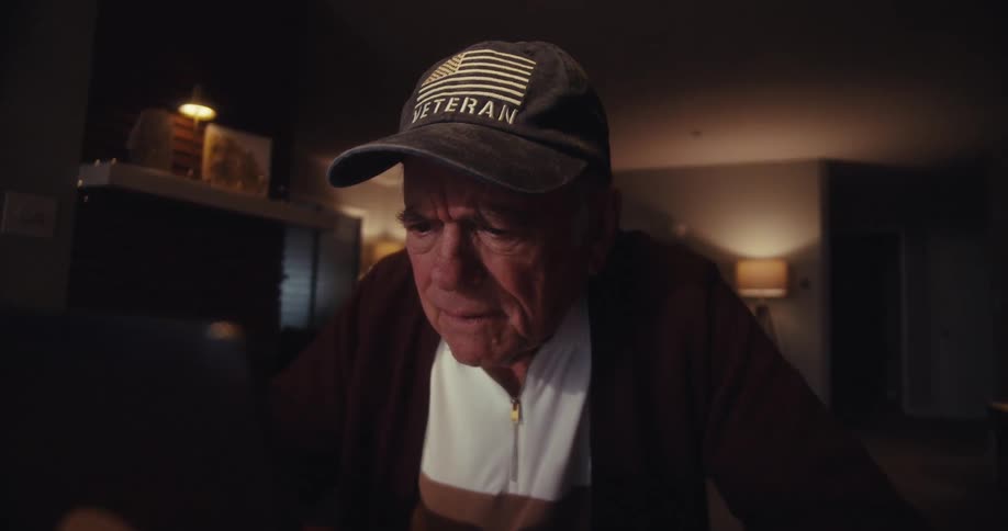 Elderly man sitting at home at night frowns then smiles while watching his computer screen