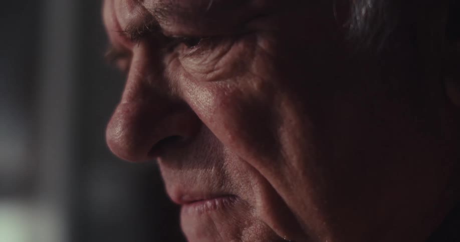 A closeup of an elderly man sitting in a dimly lit living room deep in thought and fully focused on his task