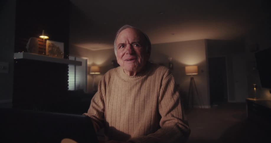 An elderly man sits at a table in a dimly lit living room smiling as he becomes lost in thought while working on his laptop