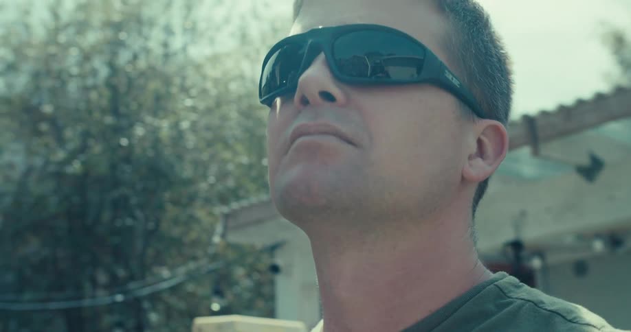 A man wearing sunglasses stands outdoors on a sunny day with buildings nearby and trees surrounding the area