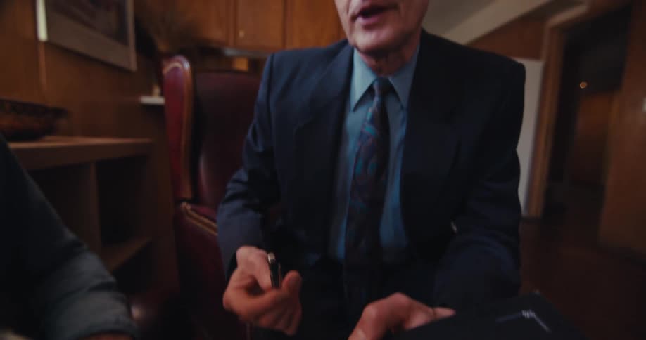 A man in a suit talks to an elderly man wearing glasses showing him a menu in his living room