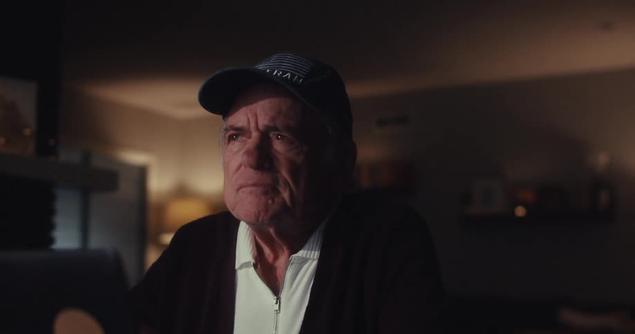 An elderly man sits alone in his living room looking sad and wearing a baseball hat