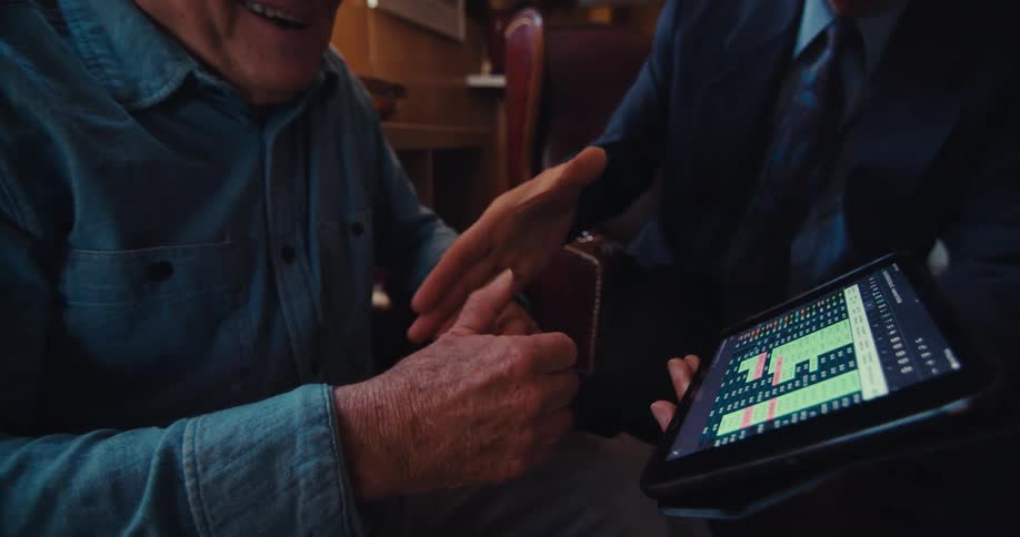 An elderly man shakes hands with a businessman who is presenting a financial chart on a tablet in a home setting