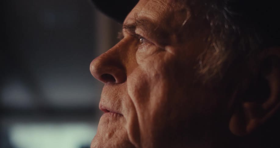 An elderly man sits in his living room looking sad then puts on a smile while wearing a hat