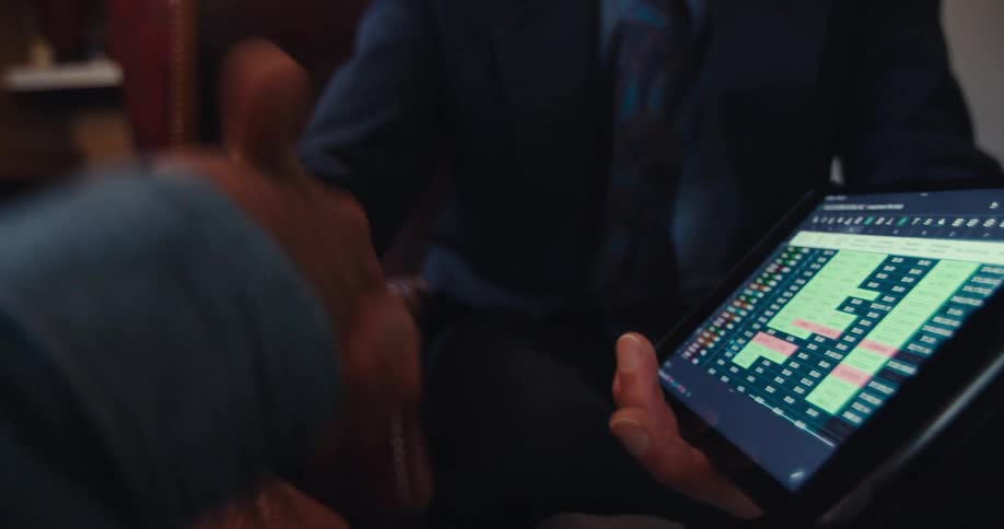 An elderly man shakes hands with a businessman who is presenting a financial chart on a tablet in a home setting