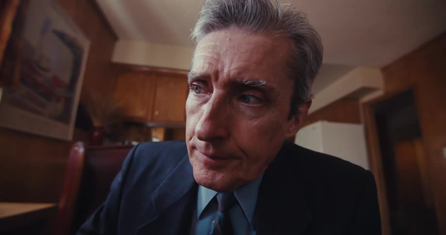 A person in a suit and tie sits in a room furnished with wooden cabinets a refrigerator and a framed painting on the wall
