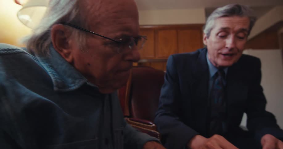 A man in a suit holds a pen and explains something to an elderly man on a tablet as they both sit in the living room
