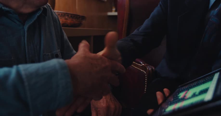 A man in a suit shows an elderly man an advertisement on a tablet pc as they both sit in the living room