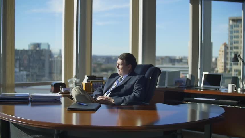 A man in a business suit sits behind a desk staring out at the skyline