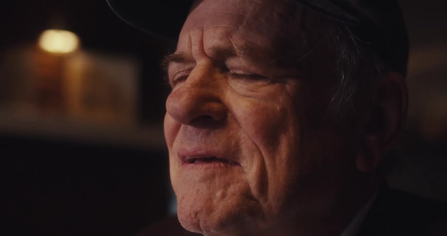 An elderly man wearing a hat sits down looking sad and then gradually changes from sad to happy